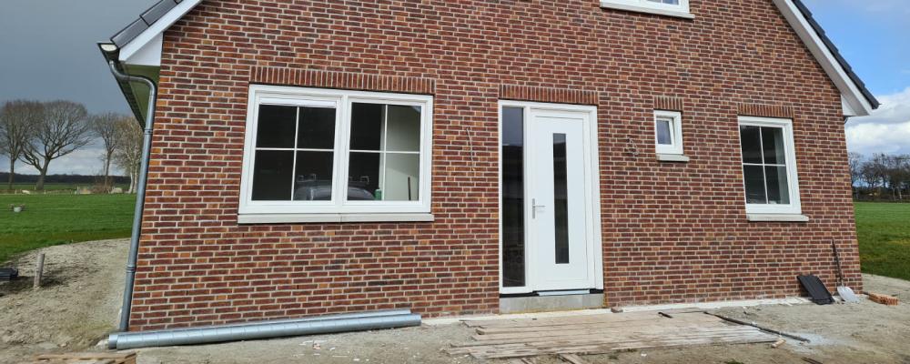 Nieuwbouwwoning met roodbruine bakstenen gevel, witte kozijnen en een zwarte pannendak, omringd door een landelijk landschap.