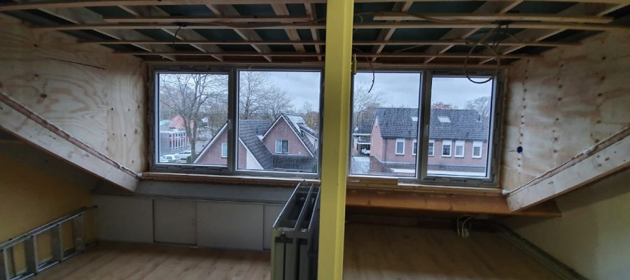 Het interieur van een zolder met een nieuwe prefab dakkapel, met zicht op omliggende huizen door de ramen.